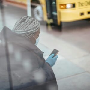 Through glass wall side view of African American female doctor using cellphone on pavement against bus on urban road